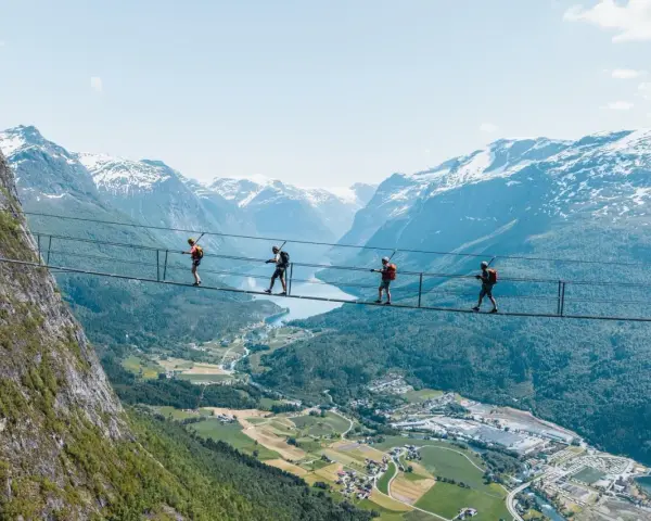 Fyra personer går på en hängbro vid Loen via ferrata, med en fantastisk utsikt över fjord och snöklädda berg.