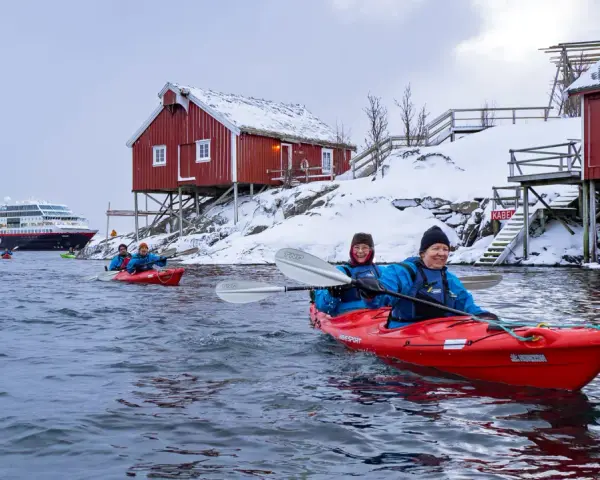 Glada paddlare i kajak i Lofoten, med snöklädda röda hus och ett stort fartyg i bakgrunden.