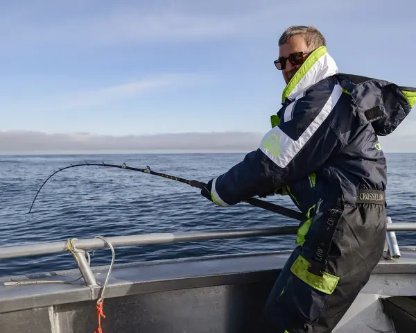 Man med solglasögon och fiskeutrustning kämpar med en böjd spö ombord på en båt. En spännande dag med fiska i Norge.