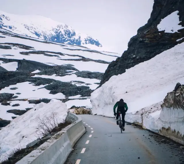 En person cyklar på en snöig väg i Norge, omgiven av höga snödrivor och berg.