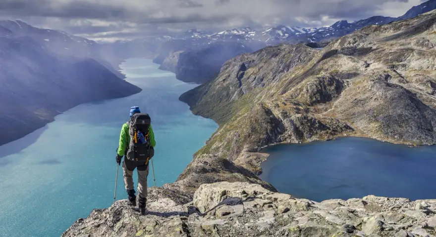 Vandrare med ryggsäck står på en klippa och blickar ut över turkosa sjöar och berg i Jotunheimen nationalpark.