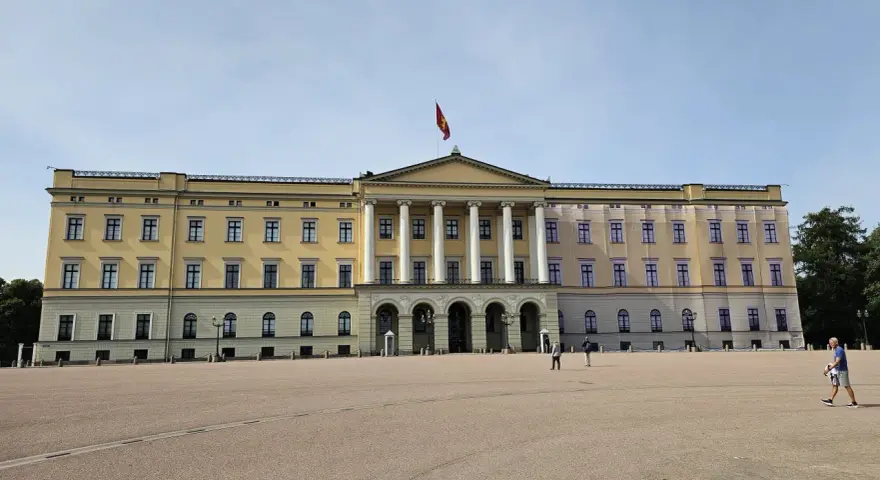Det gula **Oslo slott** med sin flagga reser sig stolt mot en klarblå himmel. En stor öppen plats framför slottet.