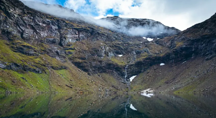 En spegelblank sjö reflekterar en dramatisk bergssida med en forsande vattenfall. Perfekt för att vandra i Abisko.