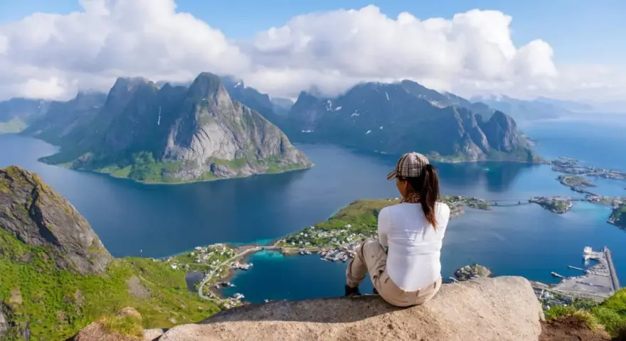 En person sitter på en klippa och blickar ut över en dramatisk kustlinje med berg och fjordar. En perfekt vy för lofoten vandring.