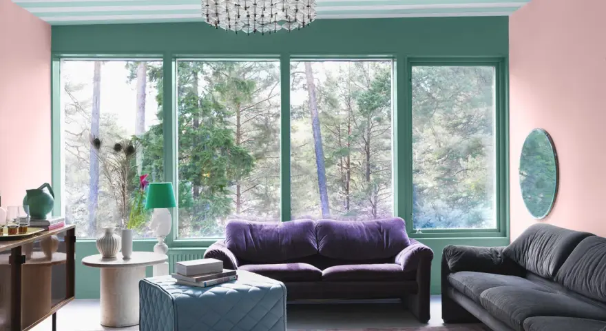 Ett vardagsrum med art deco inredning, mjuklila soffa, mörkgrå soffa, turkos pall och en stor fönsterutsikt.