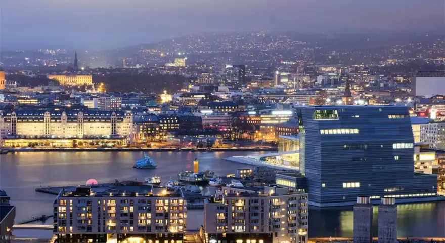 Fakta om Oslo: En stad vid vattnet med moderna byggnader och upplysta hus, som sträcker sig mot en kulle i fjärran.