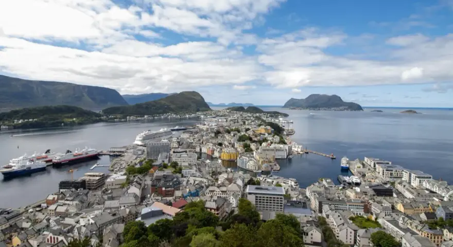 Vacker vy över Ålesund, Norge, med färgglada hus, fartyg i hamnen och berg i bakgrunden under en molnig himmel.