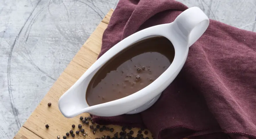 En vit såsslev fylld med mustig brunsås, bredvid en hög med svartpepparkorn. Ett perfekt brunsås recept för middagen.