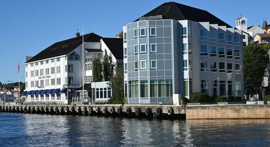 Hotellbygg vid vattnet i Arendal, Norge. En vit byggnad med blå markiser och en modern glasbyggnad står sida vid sida.