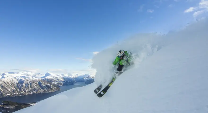 En skidåkare i grön jacka plöjer genom pudersnö med en fantastisk fjordvy i bakgrunden. Stranda ski-skidorna skär genom snön.