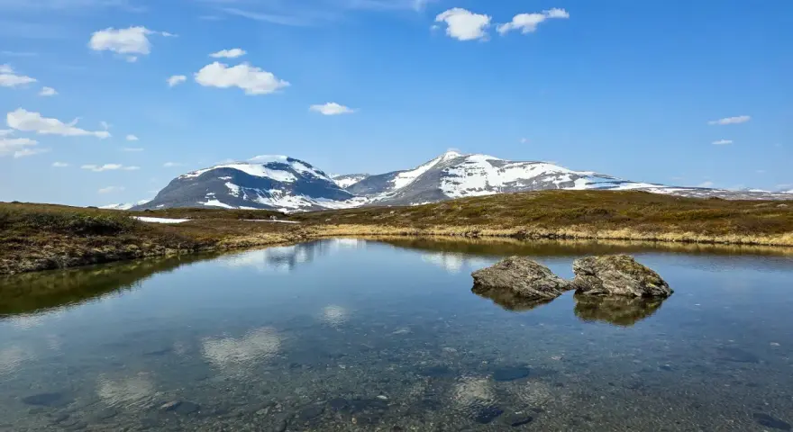 En klarblå himmel med moln speglar sig i en lugn sjö. I bakgrunden reser sig snöklädda berg, en vacker vy för ramundberget vandring.