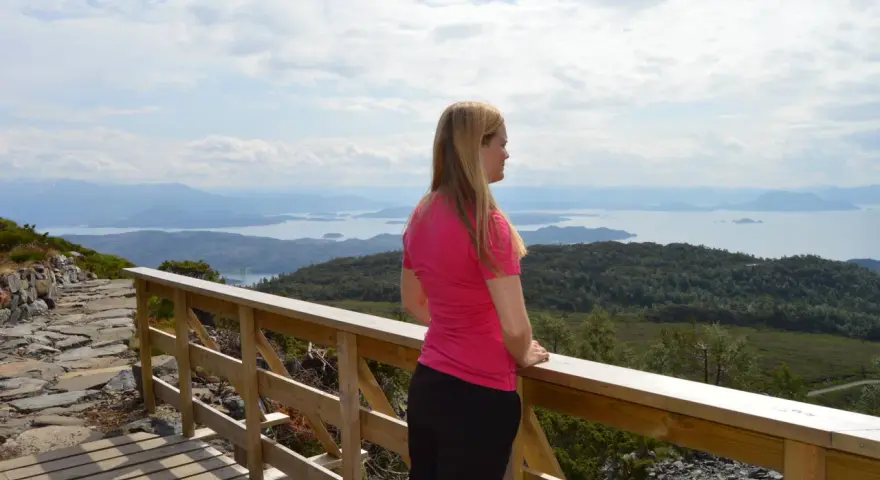Kvinna i rosa tröja njuter av utsikten över ett storslaget Norge med fjäll och fjordar.