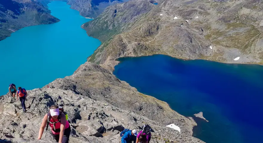 Vandrare klättrar på en stenig bergssida med turkosa och blå sjöar nedanför. En klassisk Besseggen vandring.