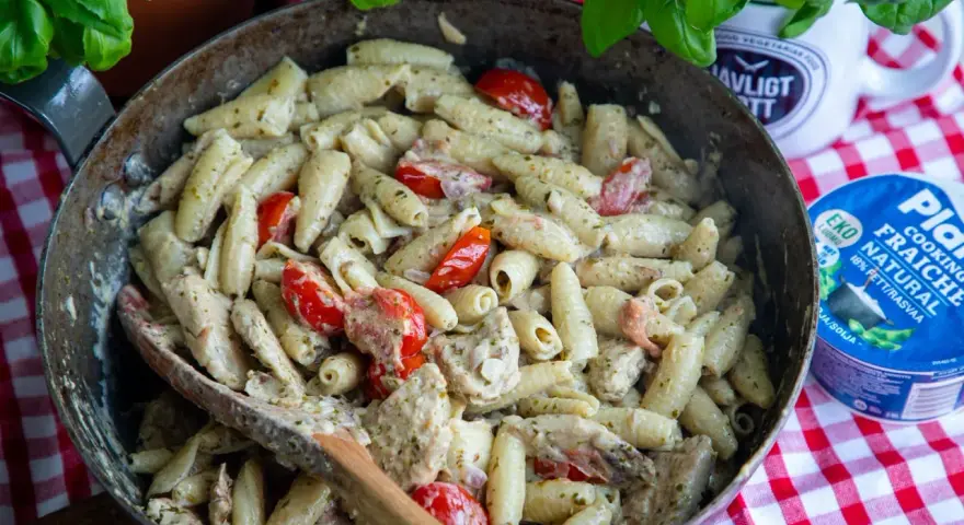 En stor panna med krämig pasta pesto, körsbärstomater och kyckling. En träsked ligger i pannan.