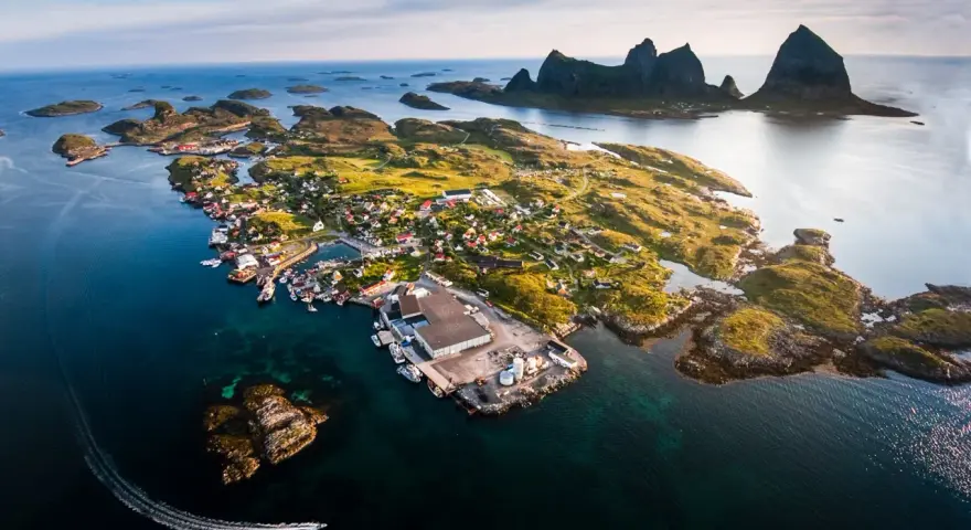 En pittoresk by på en ö i Norge, med berg i bakgrunden. En båt lämnar ett spår i vattnet. En vacker vy över traena norge.