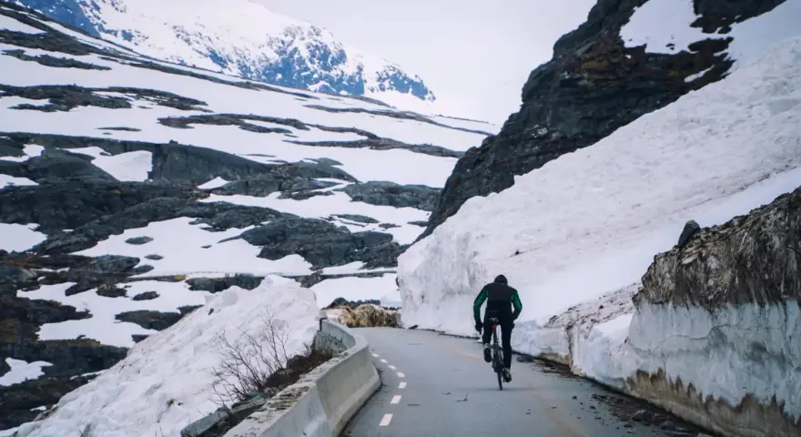 En person cyklar på en snöig väg i Norge, omgiven av höga snödrivor och berg.