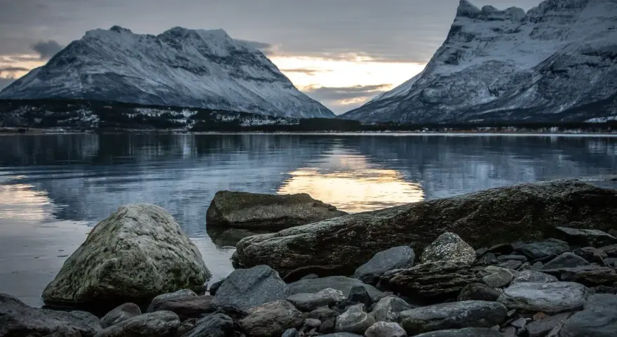 Vinterlandskap med snöklädda berg i Lyngenalperna, speglade i ett lugnt vatten. Stenar ligger vid strandkanten.