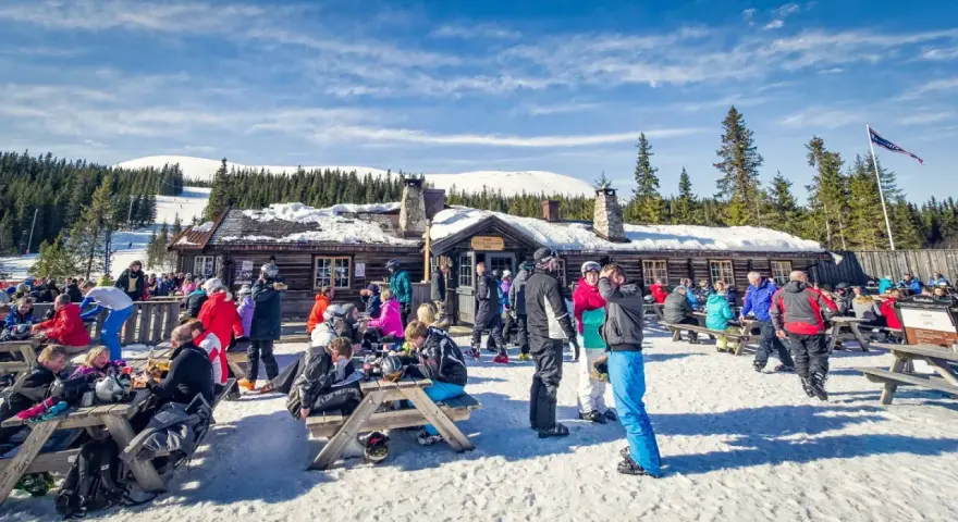 Folk njuter av afterski i Trysil vid en mysig timmerstuga med snöklädda berg och granar i bakgrunden.