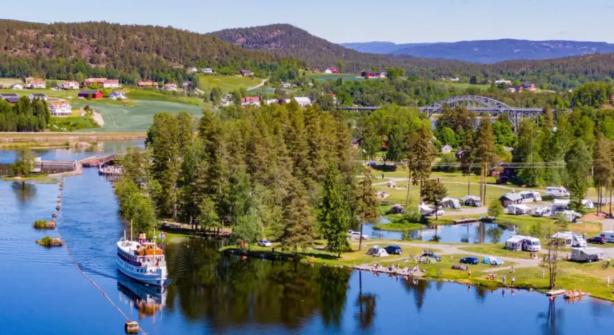 En båt på en sjö vid en campingplats i Norge. Husbilar och tält står utspridda på gräsmattor vid vattnet, omgivna av träd och kullar.