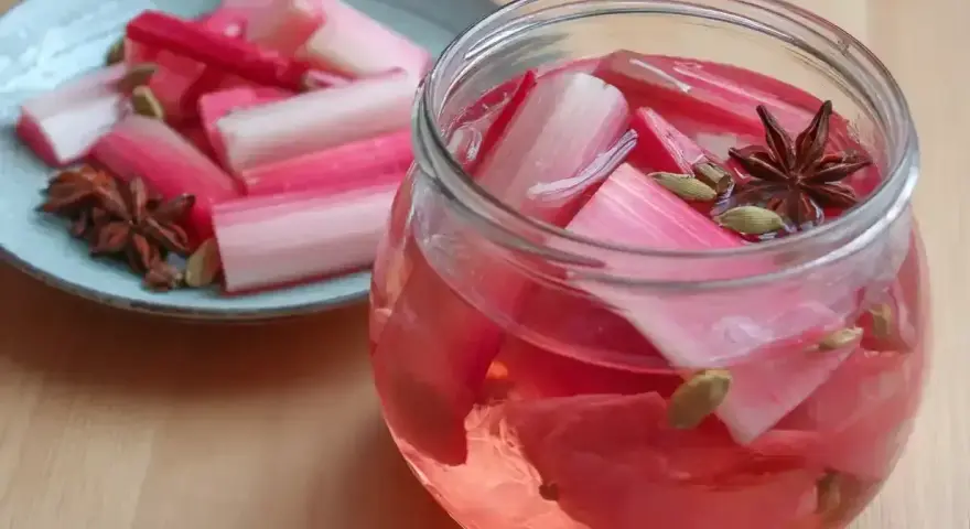 Rabarbersaft recept: färska rabarberbitar i en glasburk med kryddor som stjärnanis och kardemumma. En tallrik med rabarber och kryddor står bredvid.