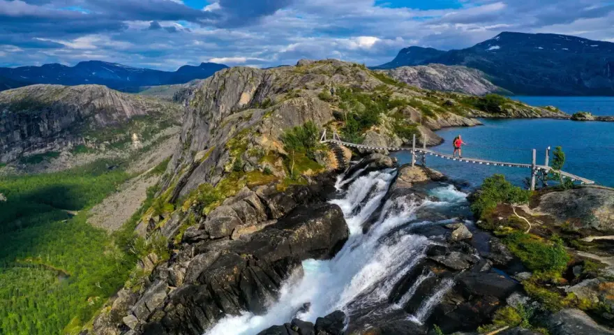 En person på en hängbro över ett vattenfall under en vandringsresa i Norge. Dramatiskt landskap med berg och en blå sjö.