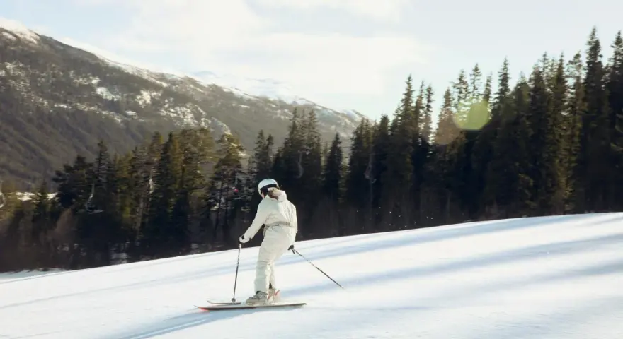 En person åker skidor utför en snöig backe, omgiven av granar och snöklädda berg. En perfekt dag på en av Sveriges skidorter.