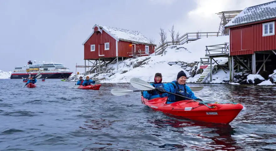 Glada paddlare i kajak i Lofoten, med snöklädda röda hus och ett stort fartyg i bakgrunden.