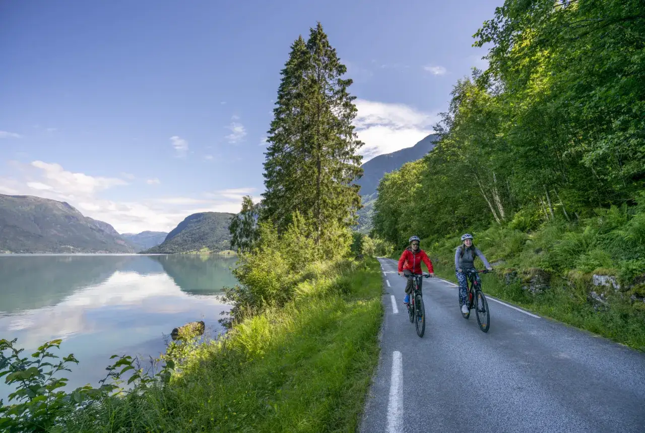 Tv&aring; personer cyklar l&auml;ngs en v&auml;g vid en spegelblank sj&ouml; i Norge. Berg och gr&ouml;nskande skog omger dem.
