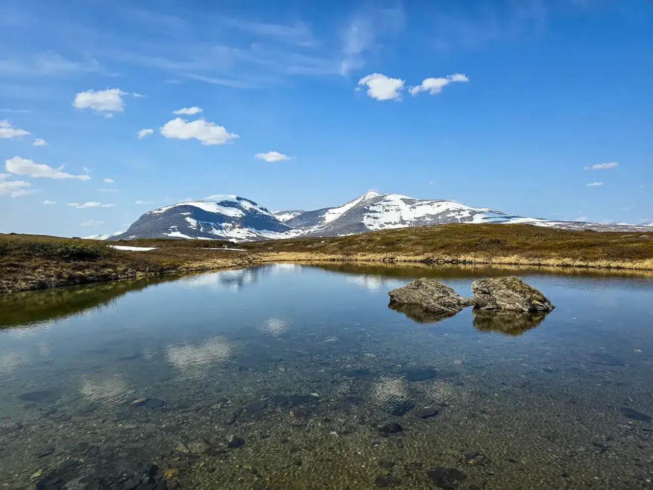 En klarblå himmel med moln speglar sig i en lugn sjö. I bakgrunden reser sig snöklädda berg, en vacker vy för ramundberget vandring.