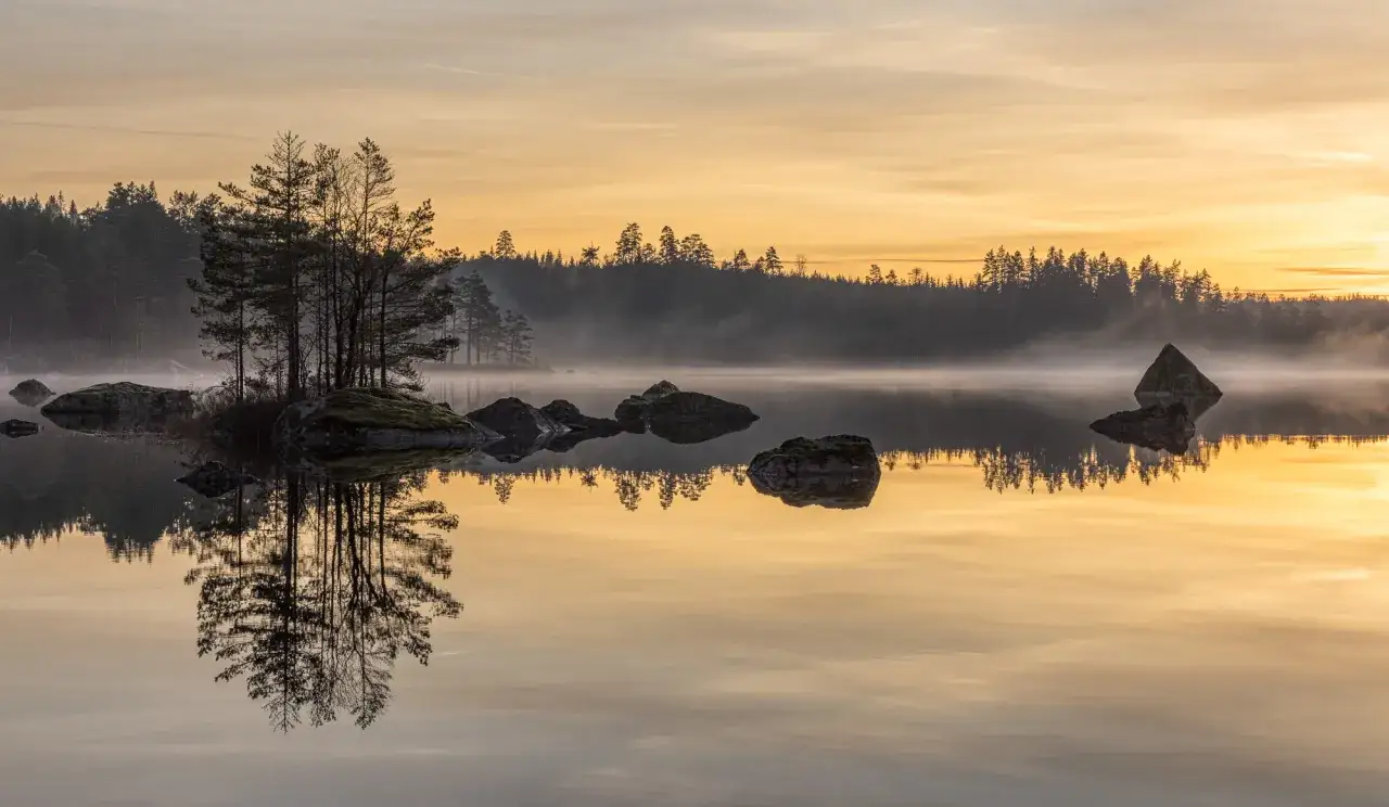 Tidig morgon vid en sj&ouml; i Kilsbergen. Tr&auml;d och stenar speglar sig i det stilla vattnet under en gyllene soluppg&aring;ng. En perfekt plats f&ouml;r kilsbergen vandring.