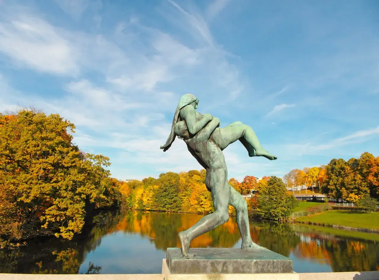 En bronsstaty av en man som bär en kvinna i Vigelandsparken, med höstlöv och en spegelblank sjö i bakgrunden.