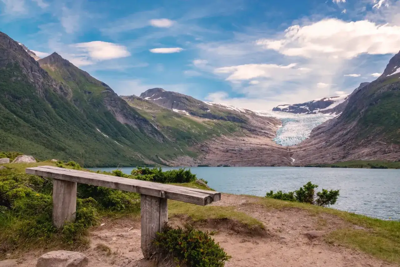 En bänk med utsikt över en glaciär och en sjö. En vy som på en Helgelandskusten karta med dramatiska berg och blå himmel.
