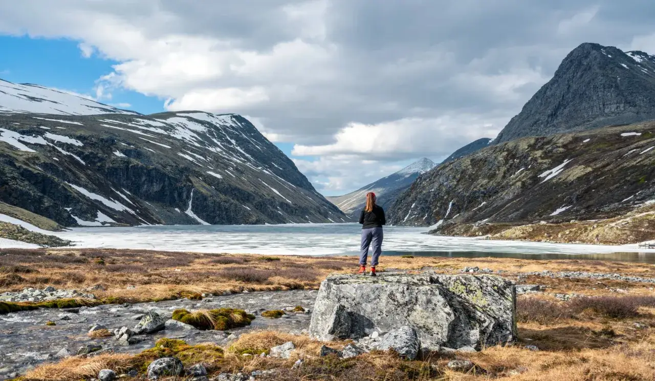 En person står vid en frusen sjö i en norsk dalgång, omgiven av snöklädda berg. En vacker vy som påminner om en triangeltur i Norge.