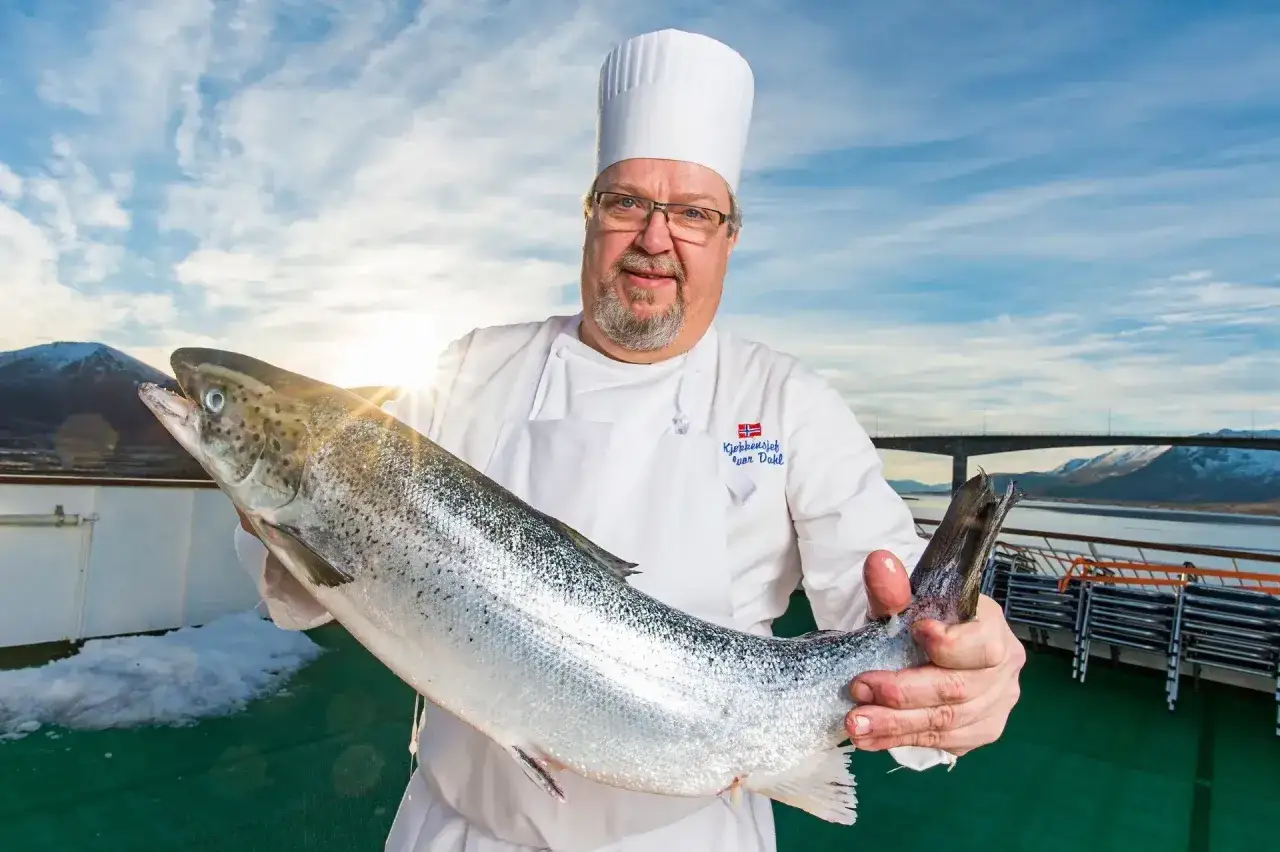 En kock i Norge stoltserar med en stor, färsk fisk. Bakom skymtar en bro och snöklädda berg under en blå himmel.