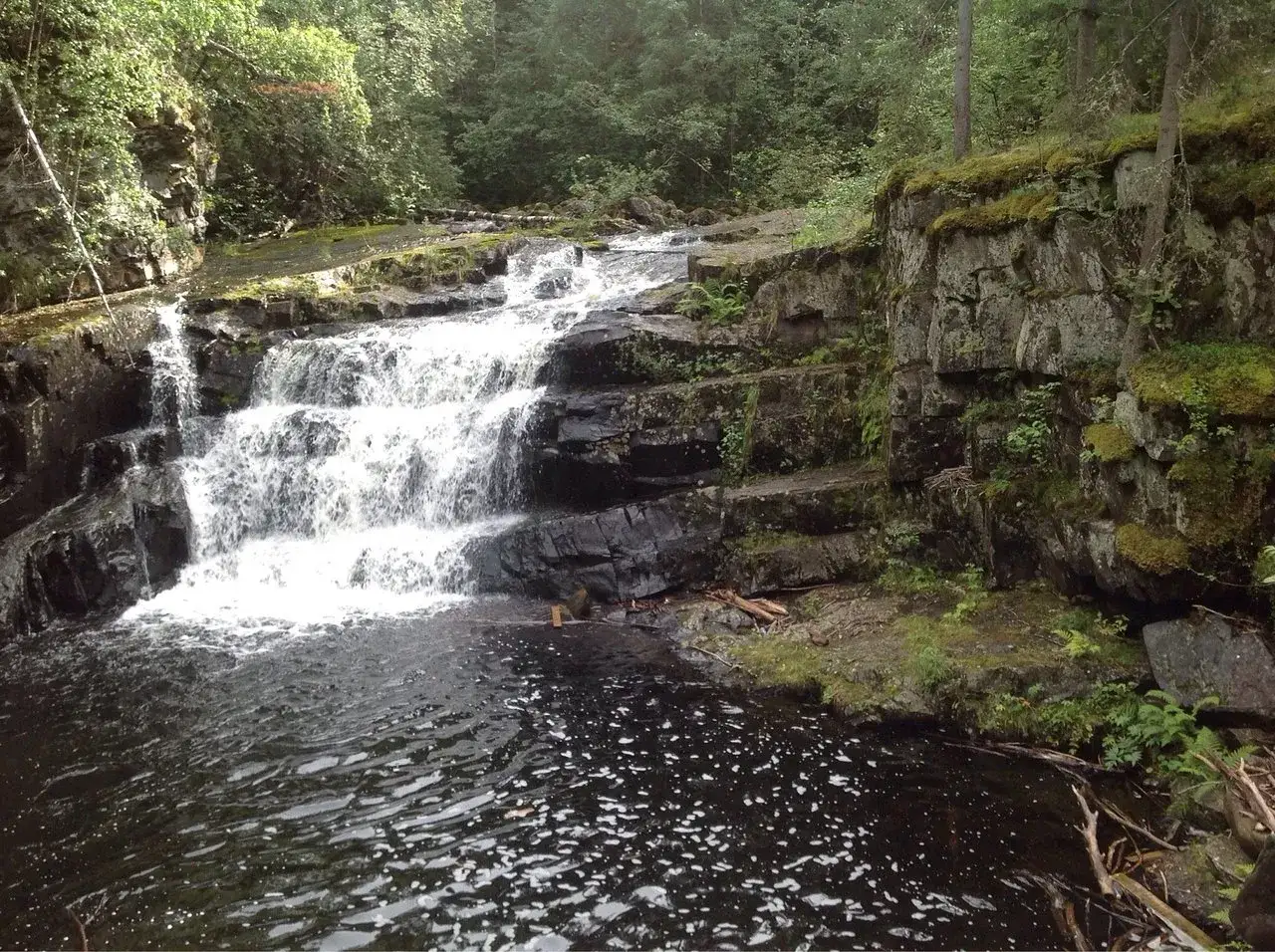 Vattenfall i en skogsmiljö nära Lillehammer, Norge. Mossbeklädda klippor och mörkt vatten.