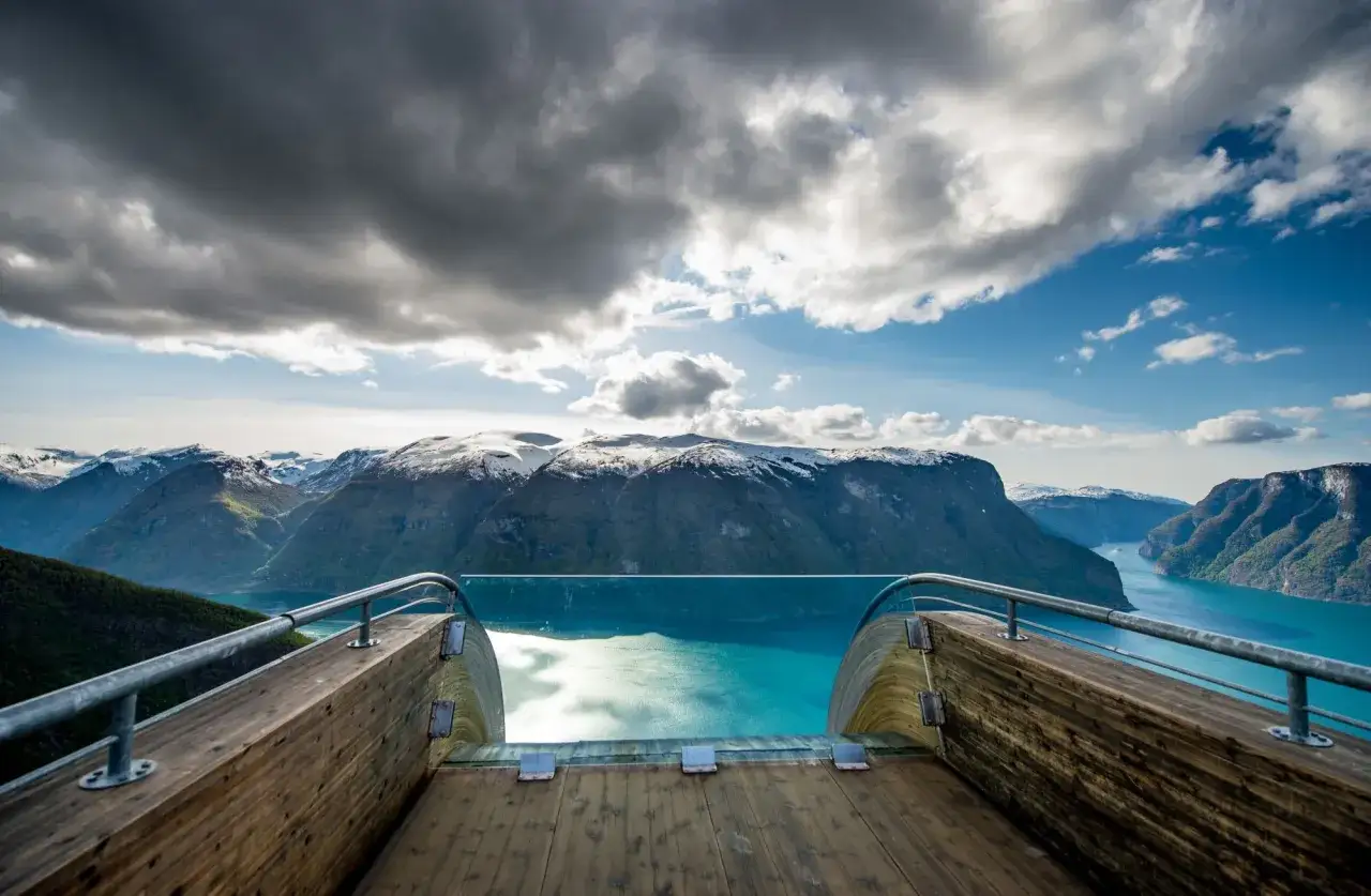 Utsikt från en utsiktsplattform i Molde, Norge, över en turkos fjord omgiven av snöklädda berg under en dramatisk molnhimmel.