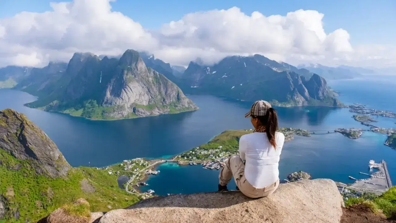 En person sitter p&aring; en klippa och blickar ut &ouml;ver en dramatisk kustlinje med berg och en by. En perfekt vy efter en h&auml;rlig lofoten vandring.
