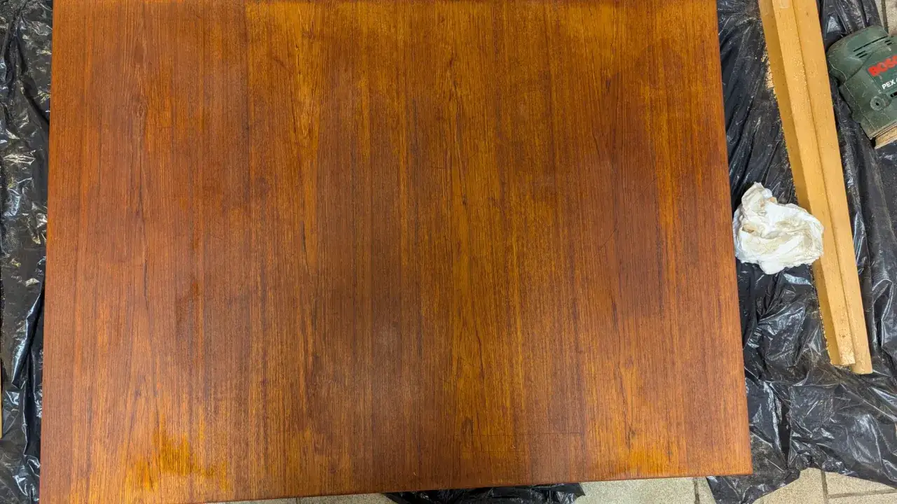 Förbereder för att slipa ett teakbord. En bit trä ligger på svart plast, bredvid en slipmaskin och en pappersbit.