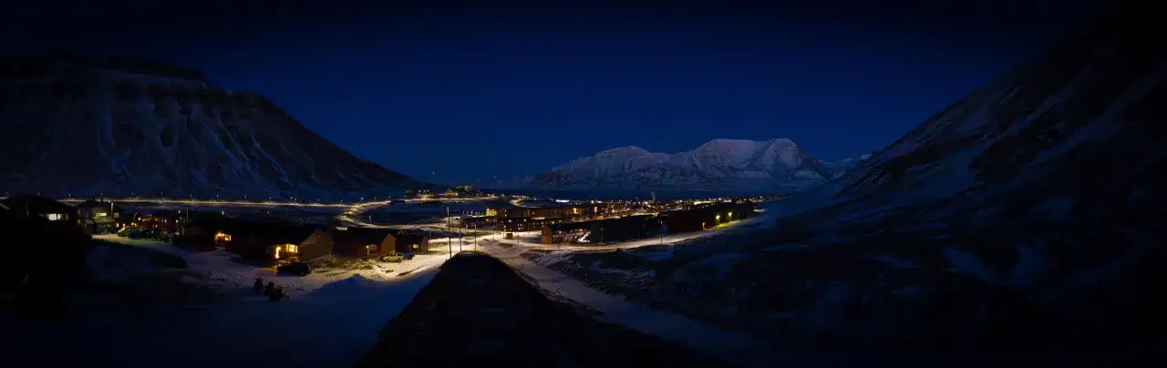 Snöklädda berg omger en upplyst by på Svalbard. En dröm att besöka Svalbard en vinternatt.