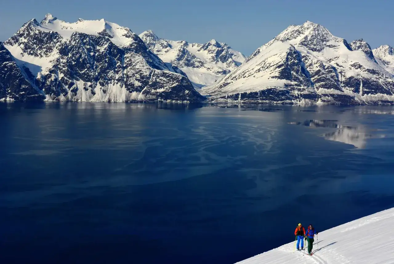 Två skidåkare på en snöig sluttning med majestätiska, snötäckta berg i Lyngenalperna som speglar sig i det mörka vattnet.