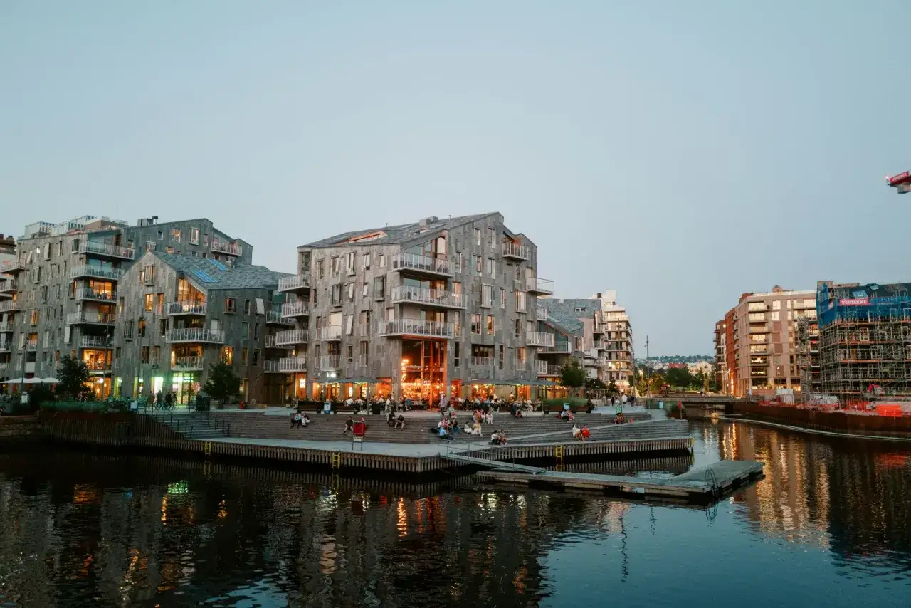 En avkopplande kväll vid vattnet i Oslo, perfekt för en weekend. Moderna byggnader speglas i vattnet medan folk njuter av kvällen.