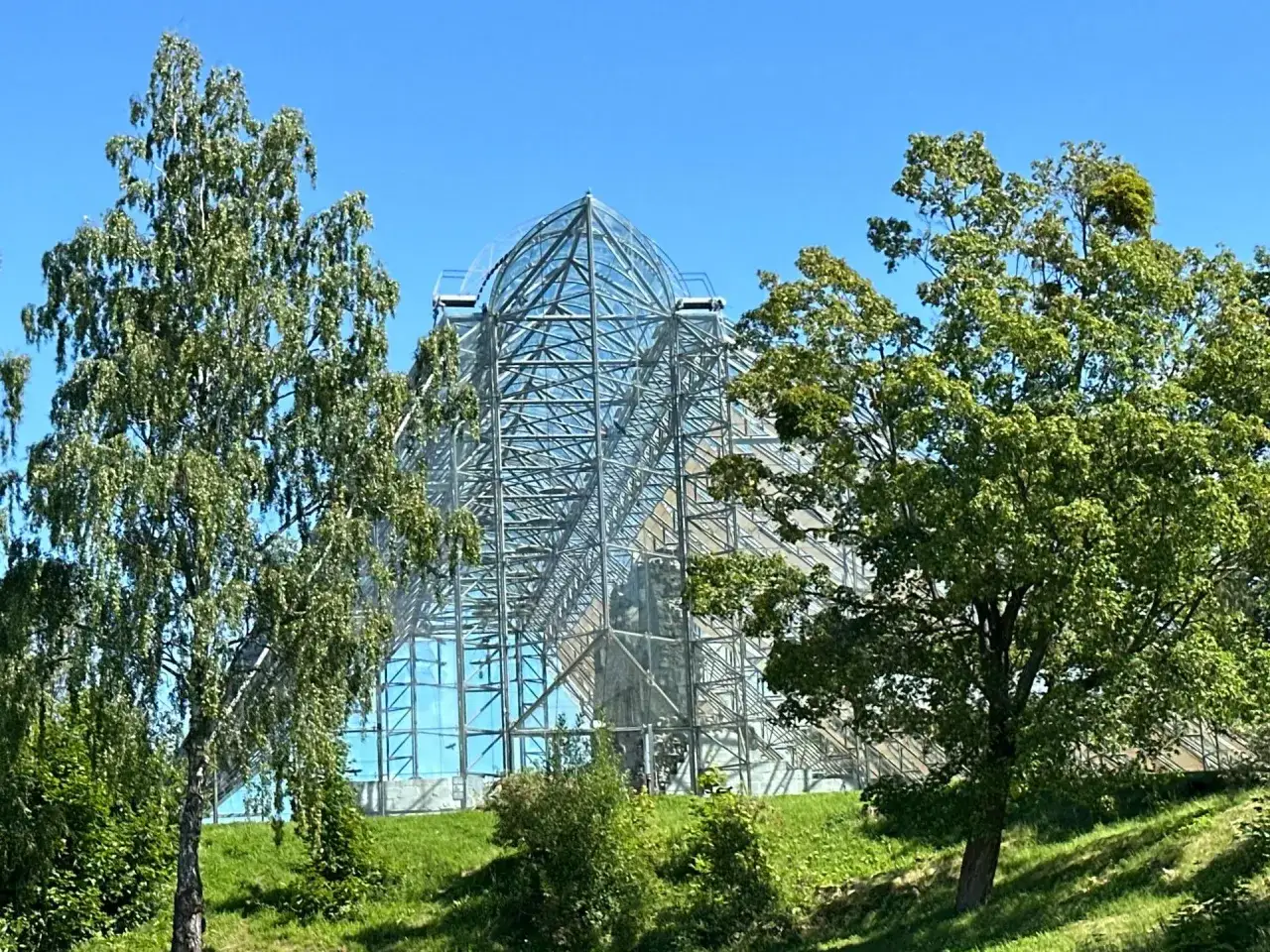 En imponerande glaspyramid reser sig mot en klarblå himmel, omgiven av grönskande träd. En vacker vy från Hamar, Norge.