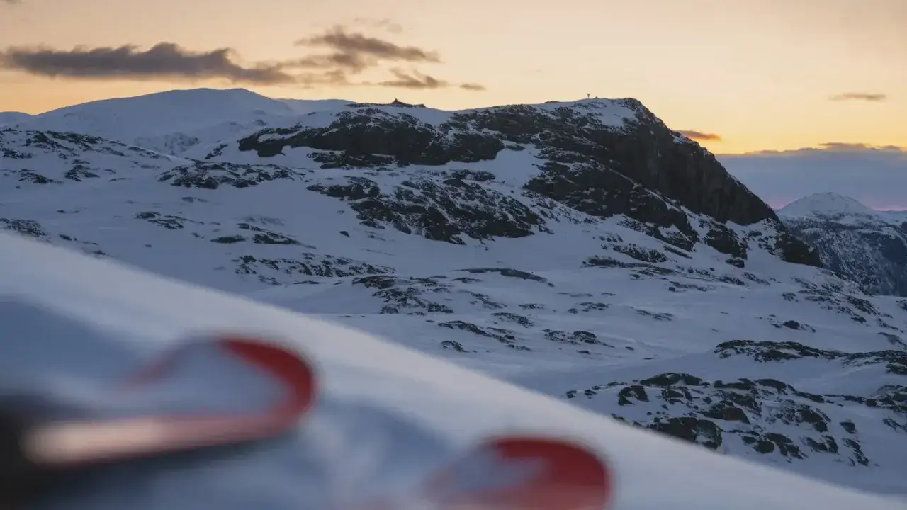 Snöklädda bergstoppar i Hemsedal, Norge, med en svag solnedgång i bakgrunden. Skidspetsar i förgrunden antyder en dag av skidåkning.