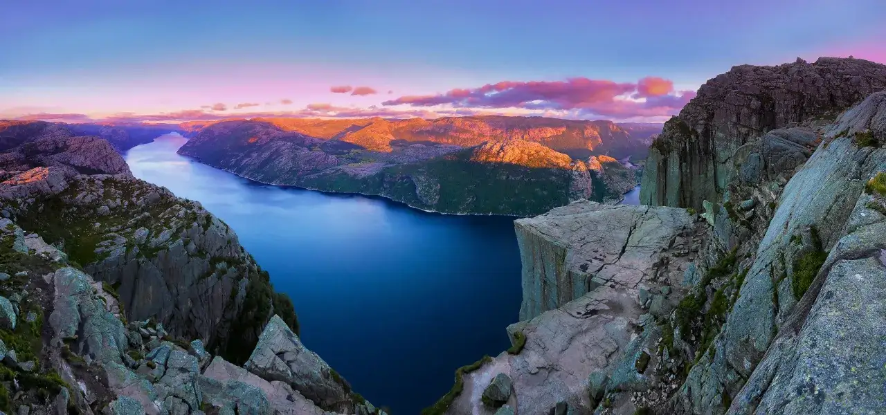Solnedg&aring;ng &ouml;ver Stavangerfjorden, med Preikestolen som sticker ut. Dramatiska klippor m&ouml;ter det djupbl&aring; vattnet under en himmel i rosa och lila toner.