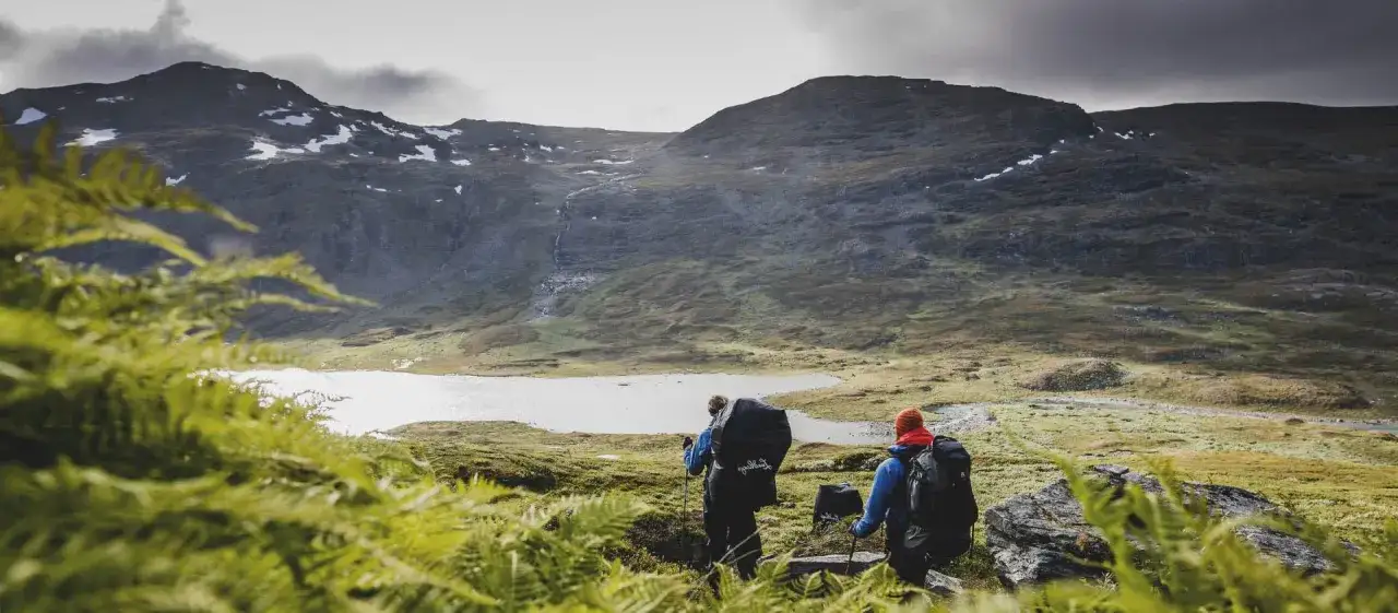 Två vandrare med ryggsäckar utforskar ett bergigt landskap med en sjö. Fina vandringsleder leder genom den vackra naturen.