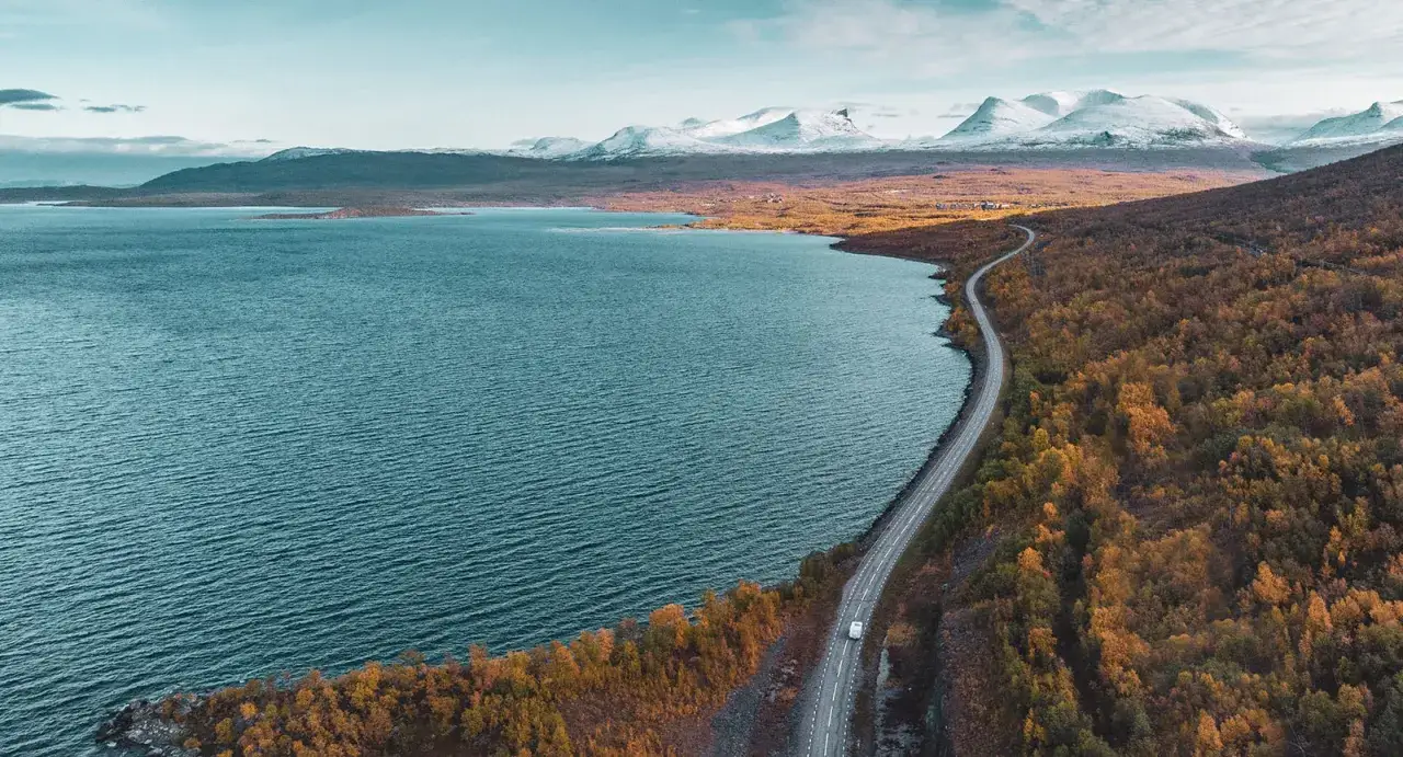 Väg slingrar sig genom höstfärgad skog längs en blå sjö, med snöklädda berg i fjärran. En resa från Narvik mot Lofoten.