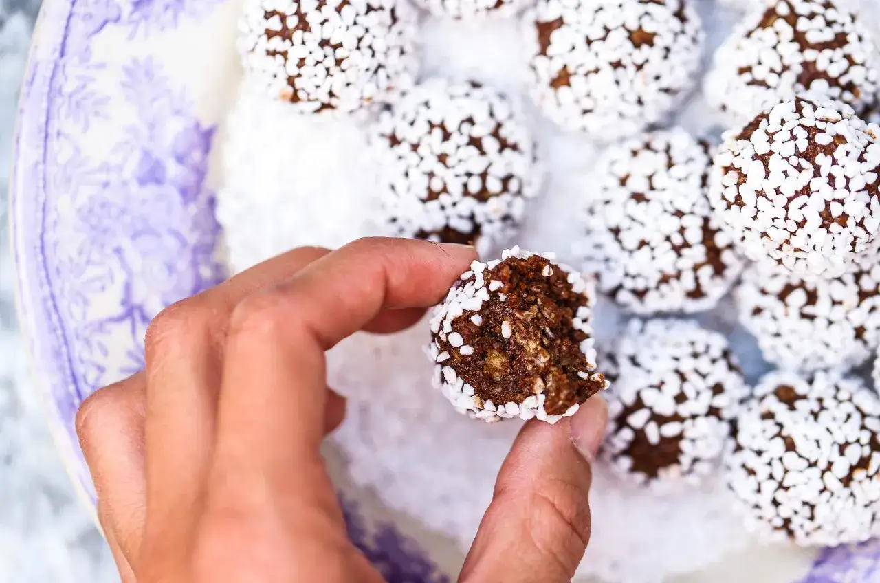 En hand h&aring;ller en bit av en chokladboll, t&auml;ckt med kokosflingor. Ett enkelt chokladbollar recept f&ouml;r hembakning.