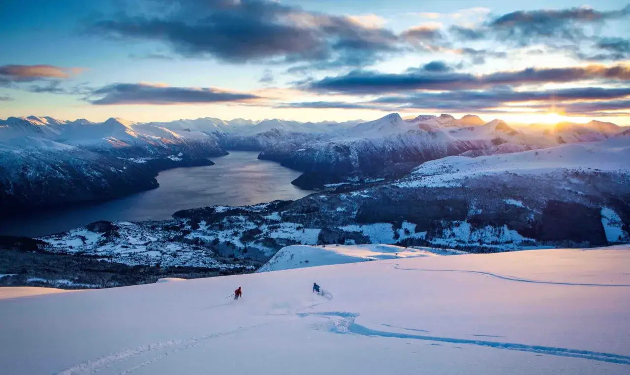 Två skidåkare njuter av solnedgången över en fjord i Norge. Vackra norge skidorter med snöklädda berg och en glittrande vattenyta.