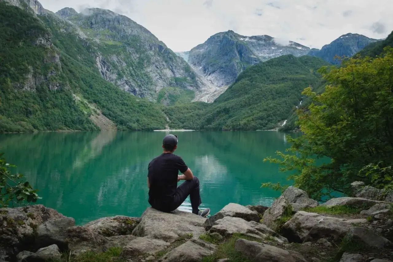 Man sitter vid en turkos sjö i Folgefonna nationalpark, omgiven av gröna berg och en glaciär i fjärran.