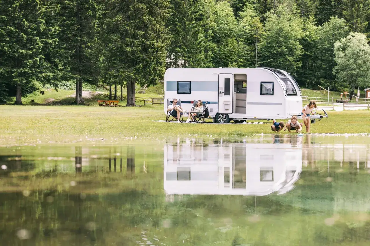 Familjen njuter av campinglivet med sin husvagn vid en sjö. Barnen leker med pappersbåtar i vattnet medan föräldrarna kopplar av i varsin stol.